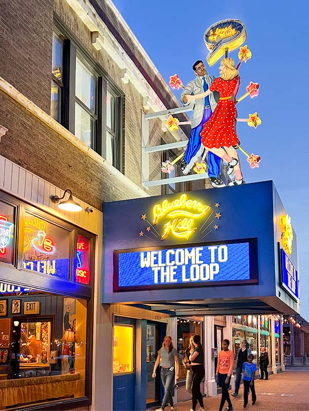 The large exterior neon sign of Blueberry Hill Restaurant & Music Club. There are two dancers on the sign with flashing neon stars. People are walking along the vibrant street.