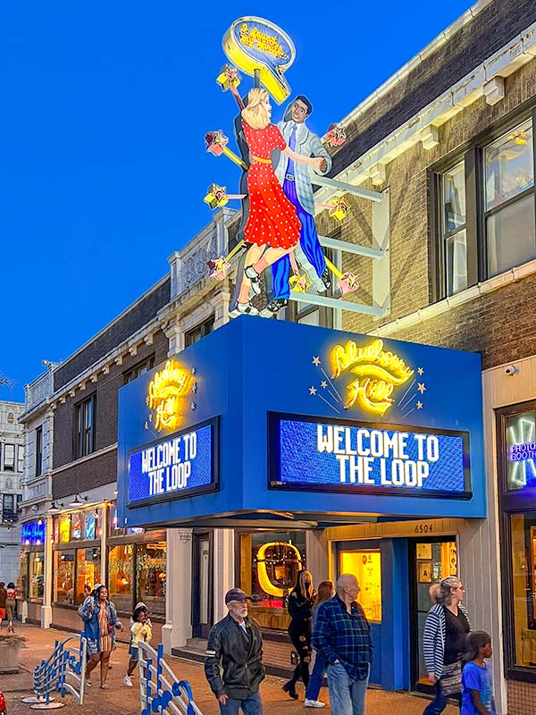 The large exterior neon sign of Blueberry Hill Restaurant & Music Club. There are two dancers on the sign with flashing neon stars. People are walking along the vibrant street.
