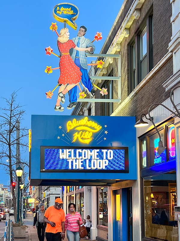 The large exterior neon sign of Blueberry Hill Restaurant & Music Club. There are two dancers on the sign with flashing neon stars. People are walking along the vibrant street.