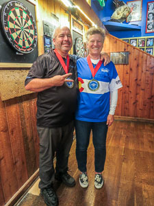 A man and woman standing in front of dart boards with their winning medals hanging around their necks. They won the 2023 Blueberry Hill Open Dart Tournament